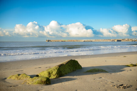 A beach with a large stone in front of the sea with a harbour arm in the background