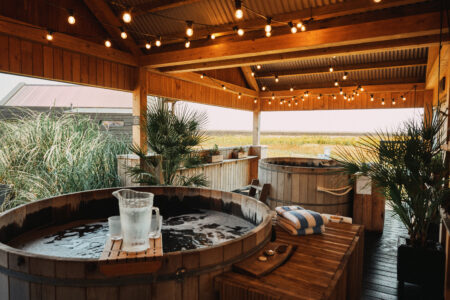 Two hot tubs sit underneath a wooden structure strung with fairy lights with a view out to grassland