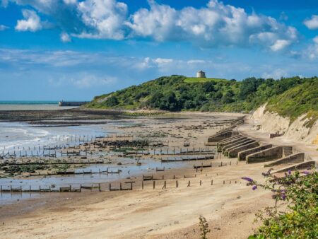 A beach with the tide out and a martello tower on a green hill in the background.