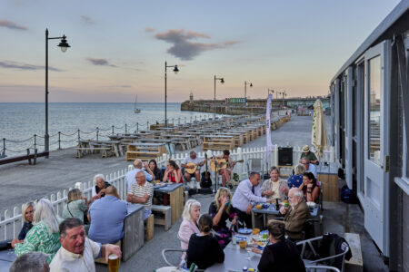 People eat and drink together in a fenced off area of a harbour arm whilst two people with guitars play.