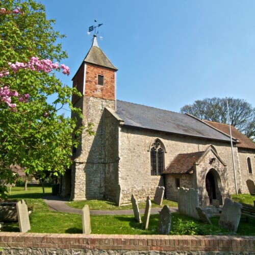 A stone church with an arched entrance sits in a graveyard with mature trees surrounding it