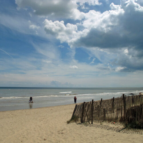 A wooden picket fence leads down towards a beach where a couple and a child play