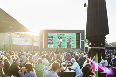 A crowd of people sit on many tables all watching a screen outside