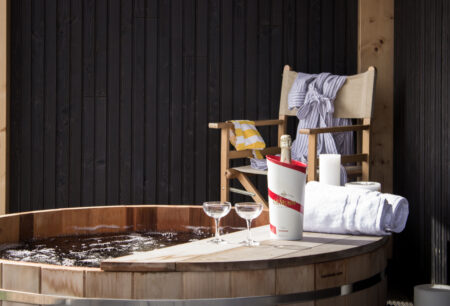 A deckchair with a dressing gown sits next to a hot tub which has a champagne bottle in a holder, two glasses and a towel on a ledge beside it