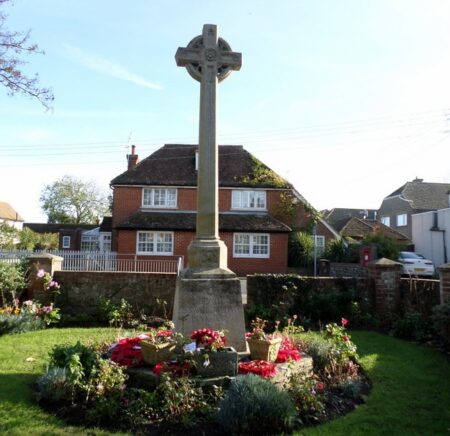 A stone cross on a plinth is surrounded by a flowerbed and then grass in a residential street.