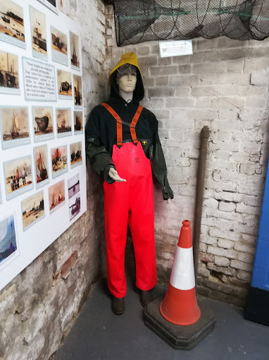 A mannequin in orange waders stands near a traffic cone and beside a board of pictures and words