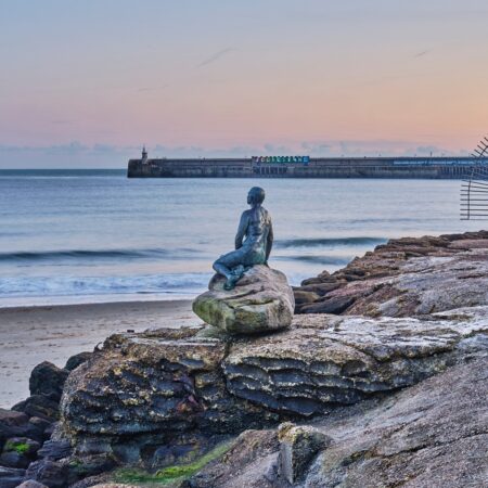 The sculpture of a mermaid sits on a rock in front of a sandy beach