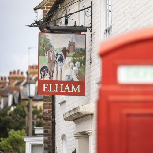 A pub style sign which says Elham with a picture of a man herding sheep and a cow. There is a red postbox in the foreground of the picture and a row of houses behind