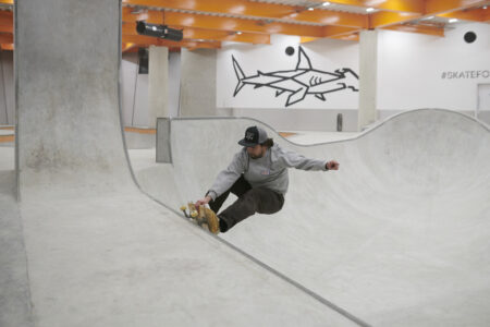 A skater does a trick in a concrete bowl with a white wall with a shark mural behind