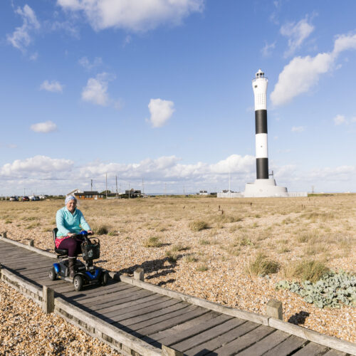 A lady uses a mobility scooter on a boardwalk with a black and white lighthouse in the background