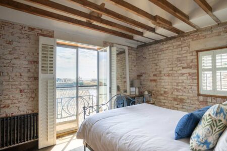 A bedroom with brick walls and beams has glass balcony doors which open to a view of rooftops